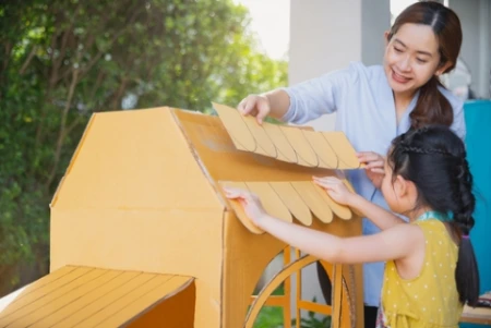 Seorang anak perempuan sedang membuat rumah-rumahan dari kardus bersama Ibu.